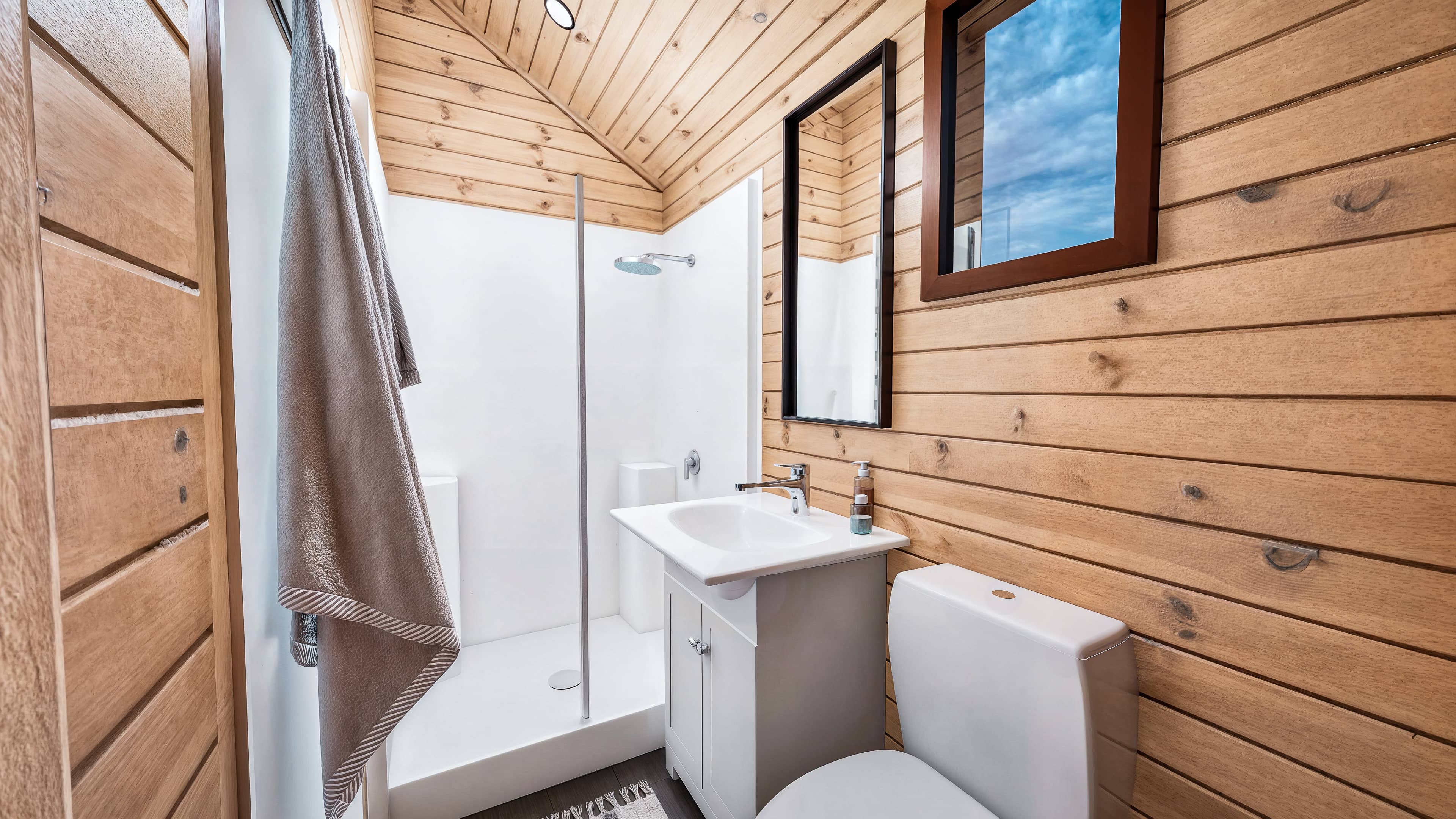 A Small wood-paneled bathroom, a shower with a gray towel, a white sink and cabinet, a mirror, a window, and a toilet.