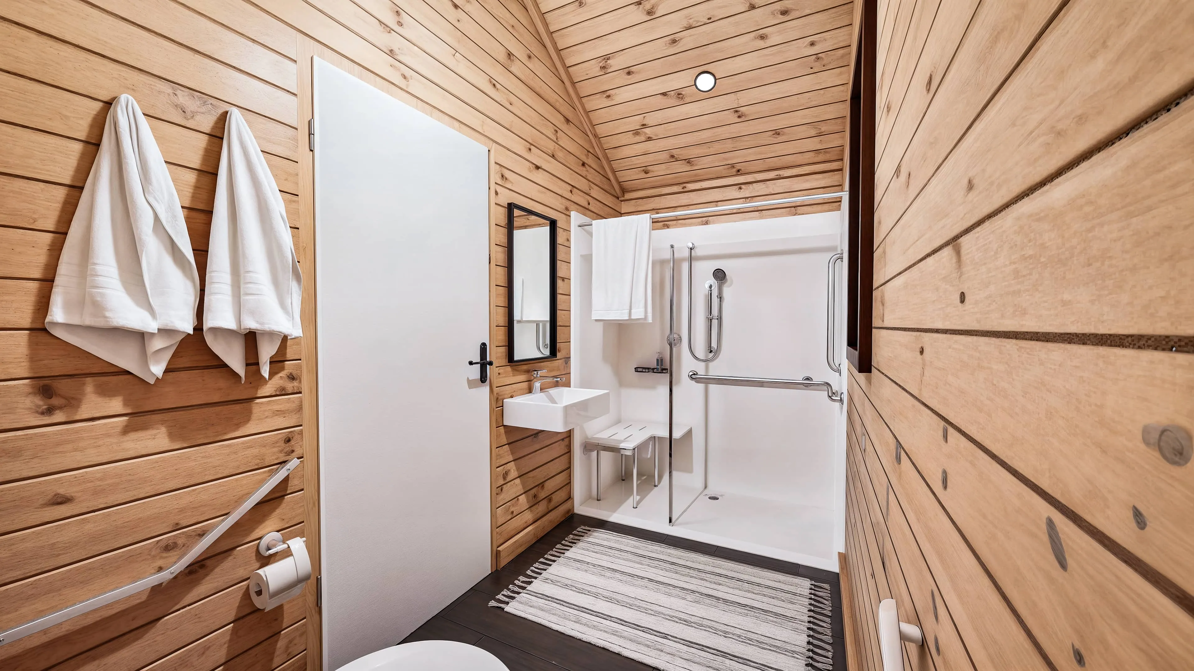 Bathroom with wood-paneled walls, accessible shower with grab bars and seat, small sink with mirror, two towels on hooks, striped rug, and a white door.