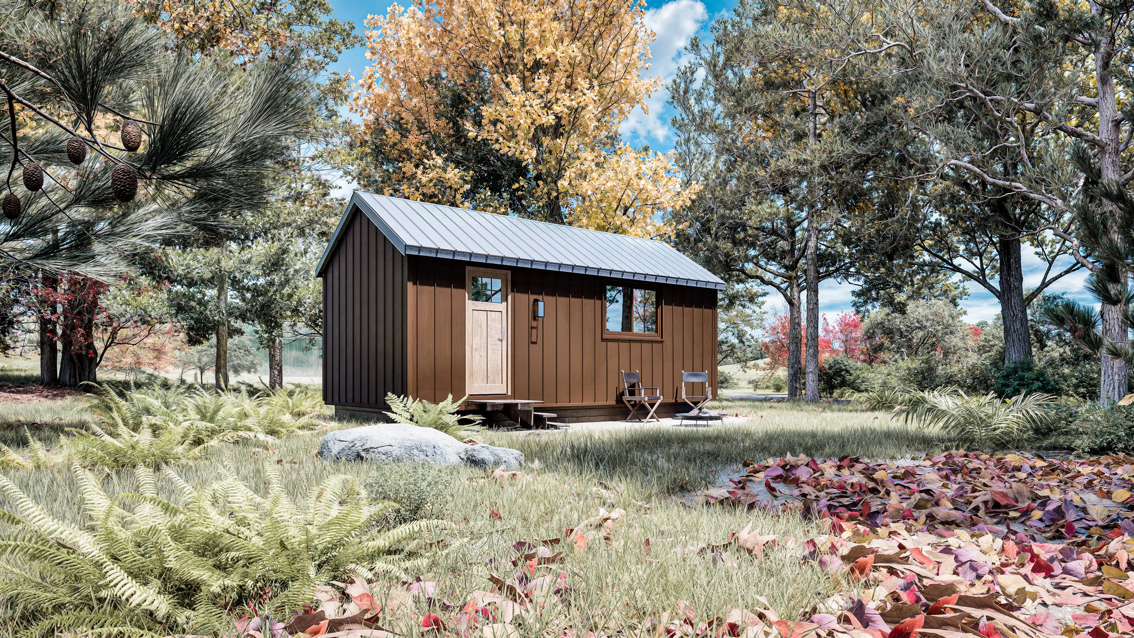 A small wooden cabin with a metal roof stands in a grassy clearing surrounded by trees, ferns, and autumn leaves. Two chairs and a table sit on the porch.