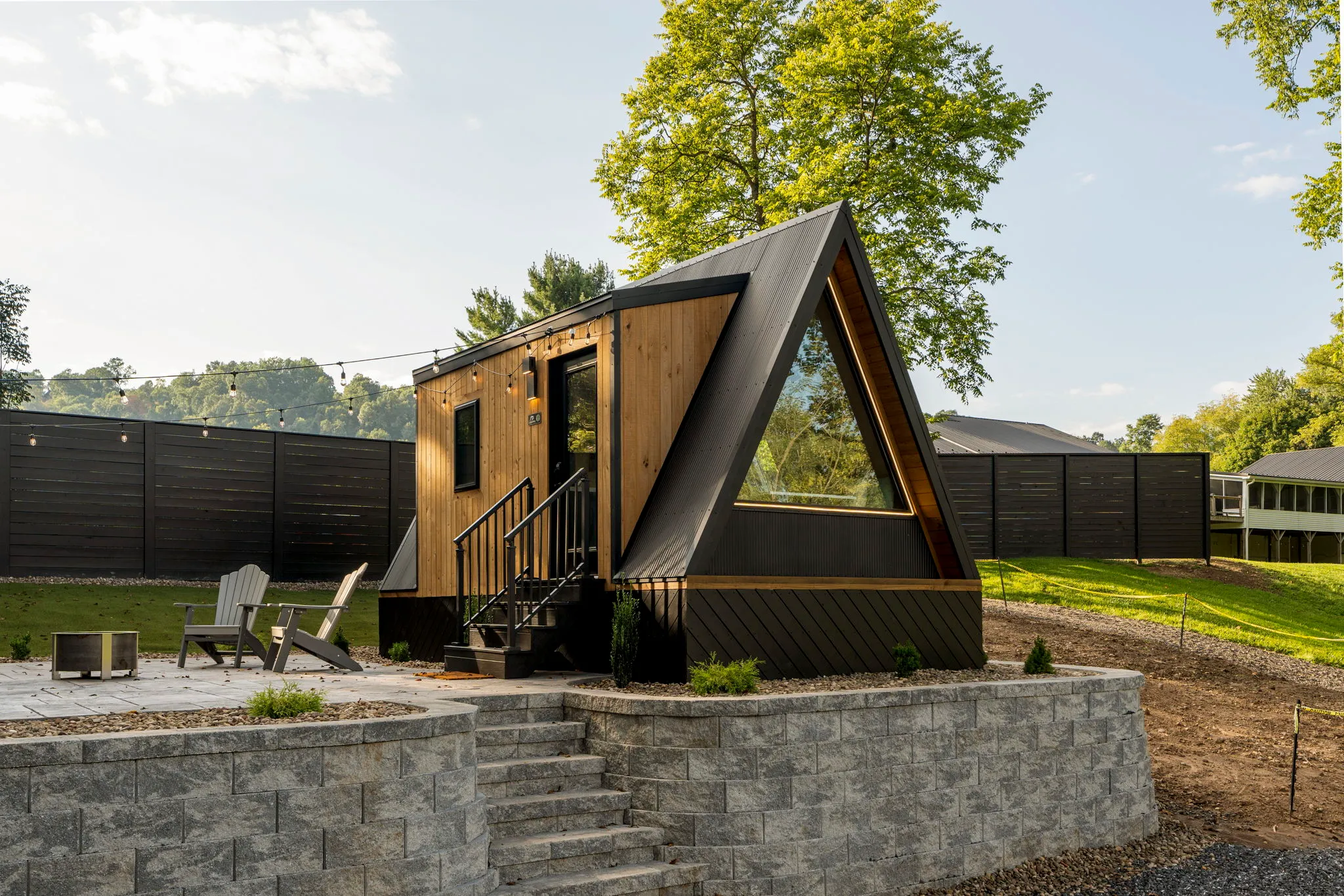 A modern A-frame tiny house with large windows sits on a raised stone patio with steps, outdoor chairs, and a fire pit, surrounded by trees and a tall privacy fence.