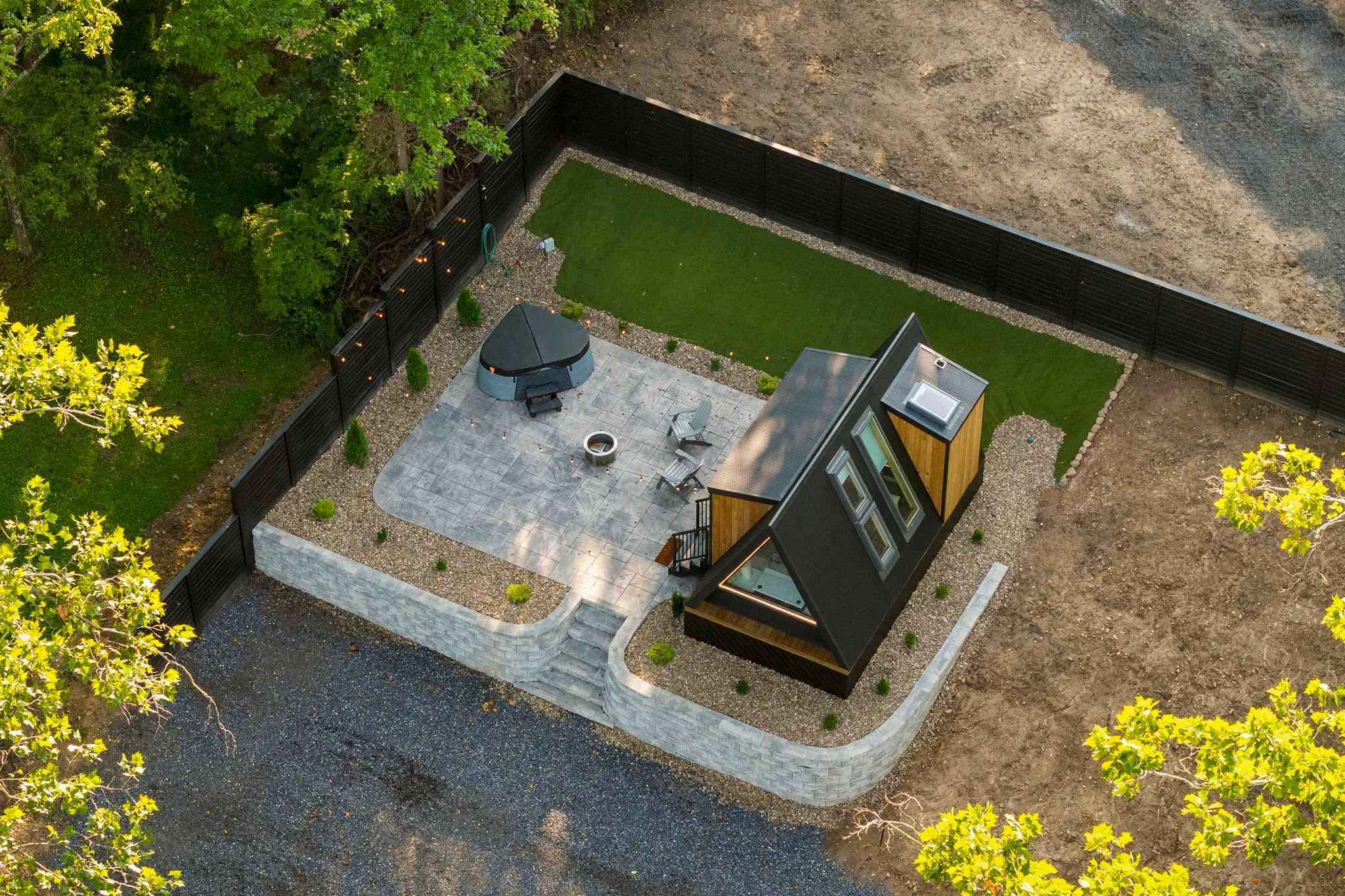 Aerial view of a small A-frame cabin with a patio, fire pit, outdoor furniture, artificial grass, and surrounding fence.