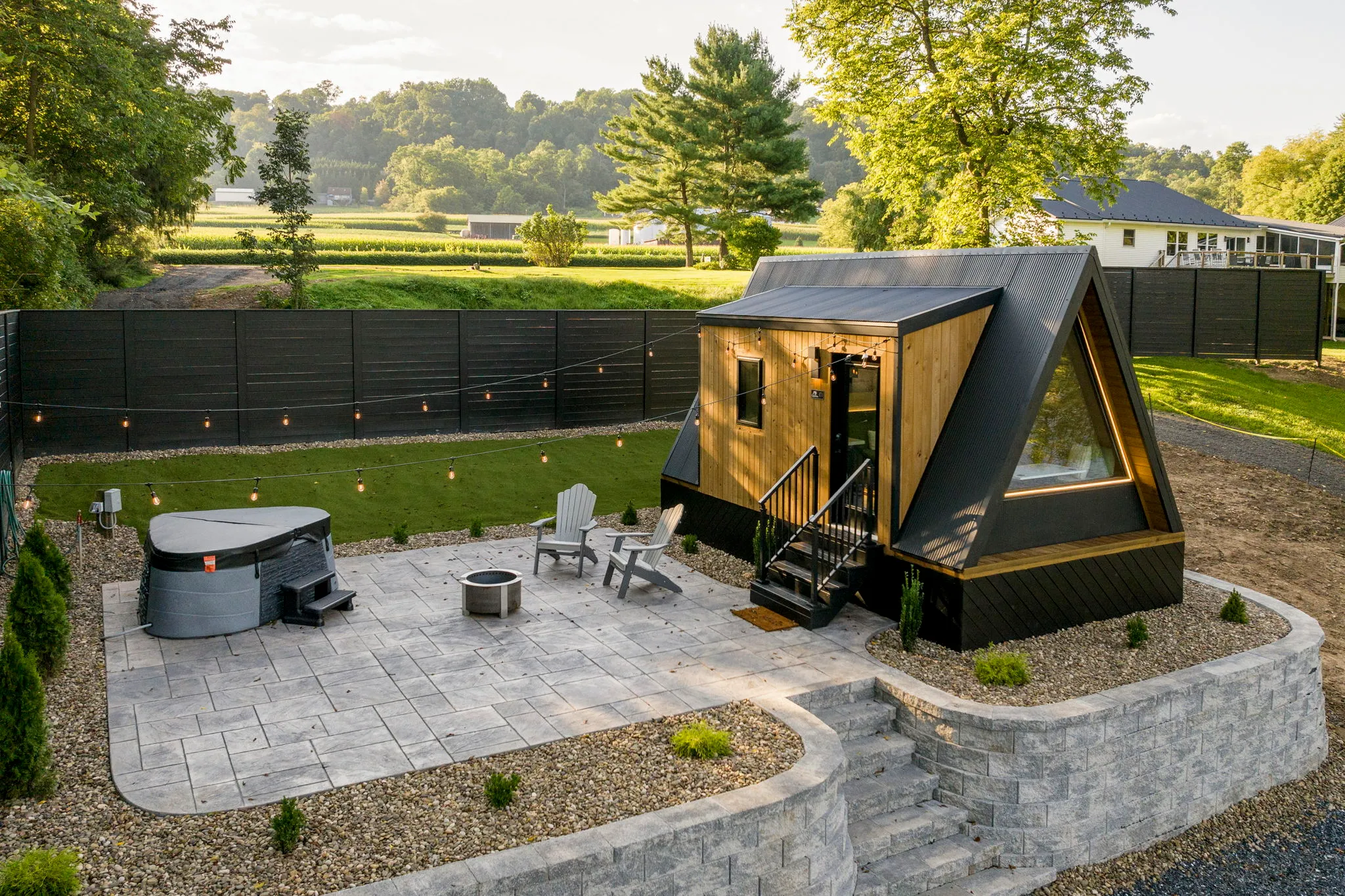 Modern A-frame backyard cabin with patio, fire pit, outdoor chairs, landscaped stone walls, and lush trees in warm evening light.