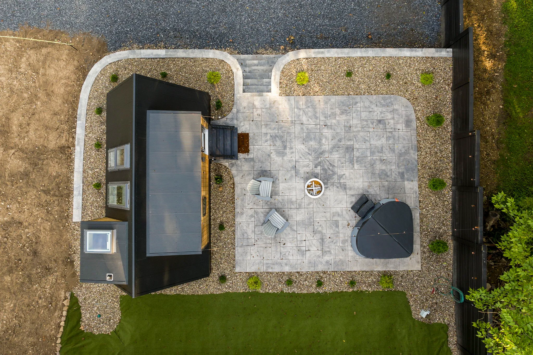 Aerial view of a modern backyard with a black-roofed home, gray stone patio, outdoor chairs, round fire pit, gravel beds, and green lawn.