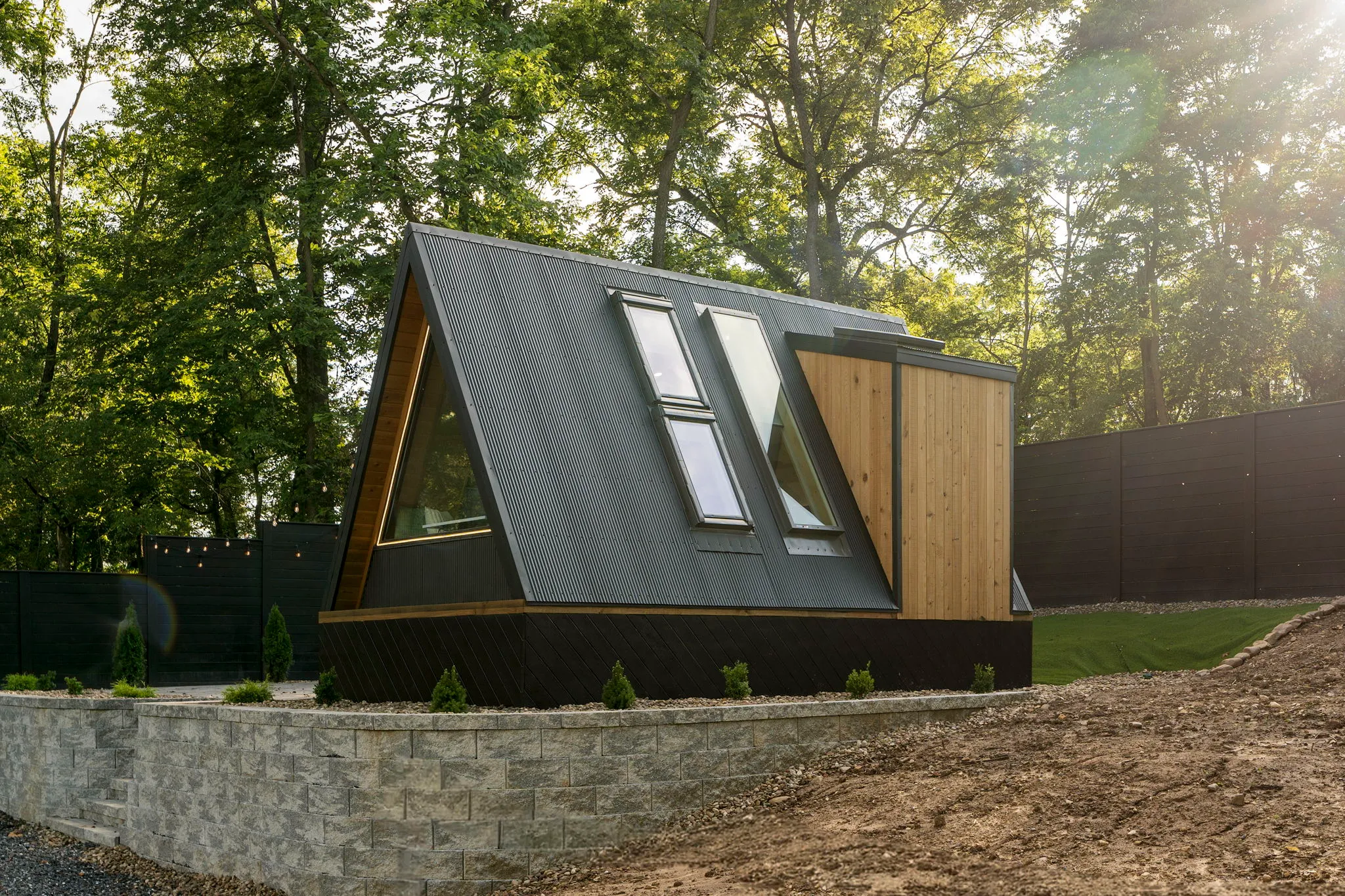 Modern A-frame cabin with dark siding, skylights, and a wooden door, nestled in sunlit woods above a stone retaining wall; serene setting.