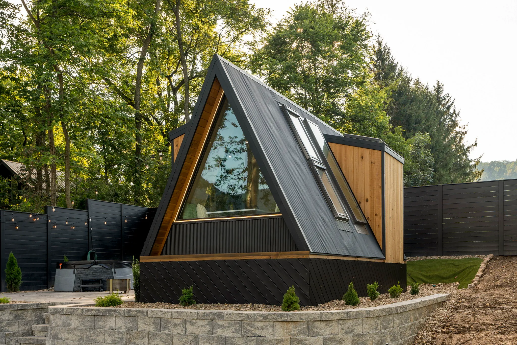 Modern A-frame cabin with black siding, warm wood trim, large windows, and a steep metal roof, set in a peaceful leafy woodland garden.