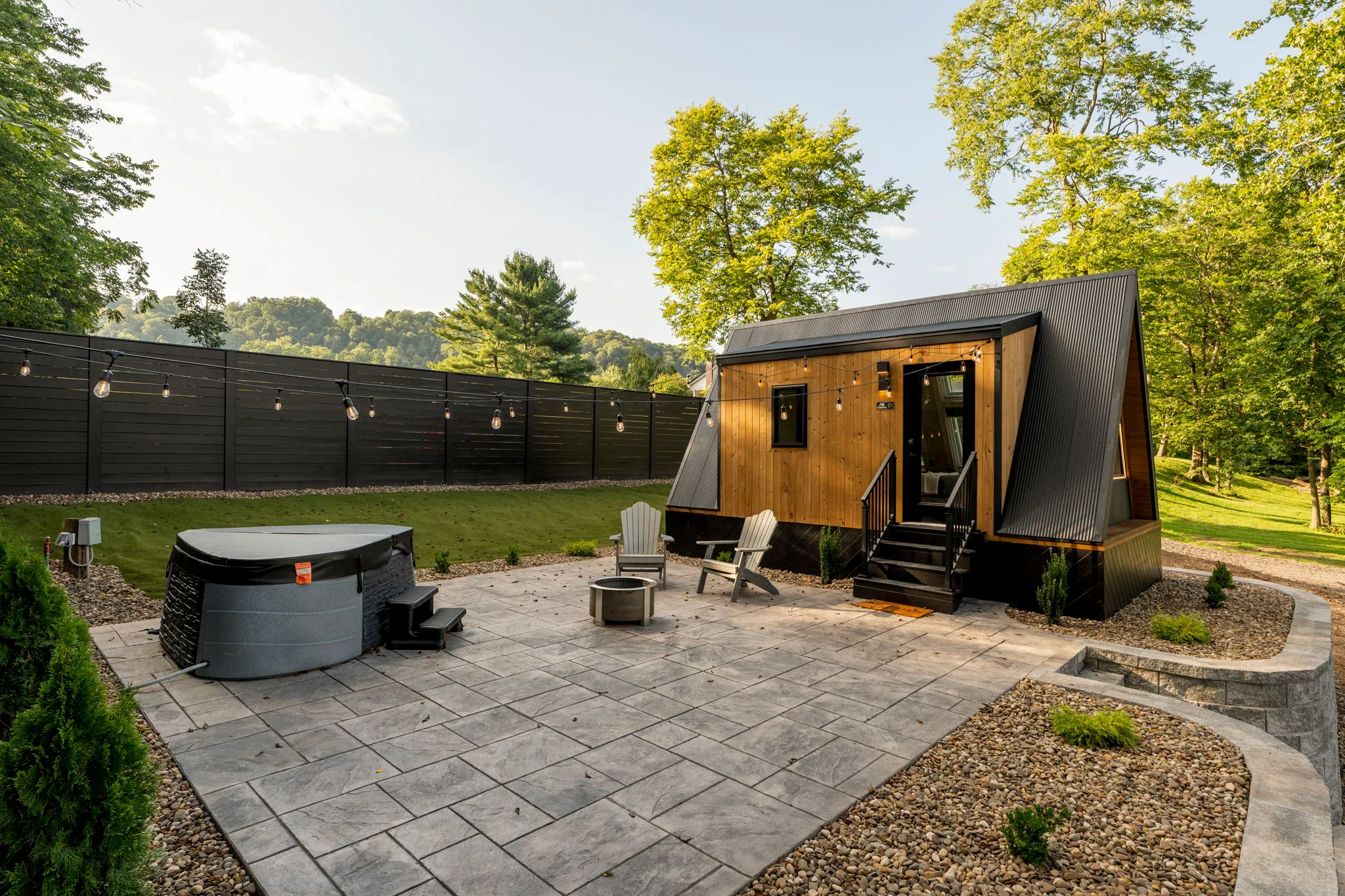 Modern tiny cabin with wood siding and black roof on a paved patio, hot tub, chairs, and fire pit, surrounded by peaceful green woods.
