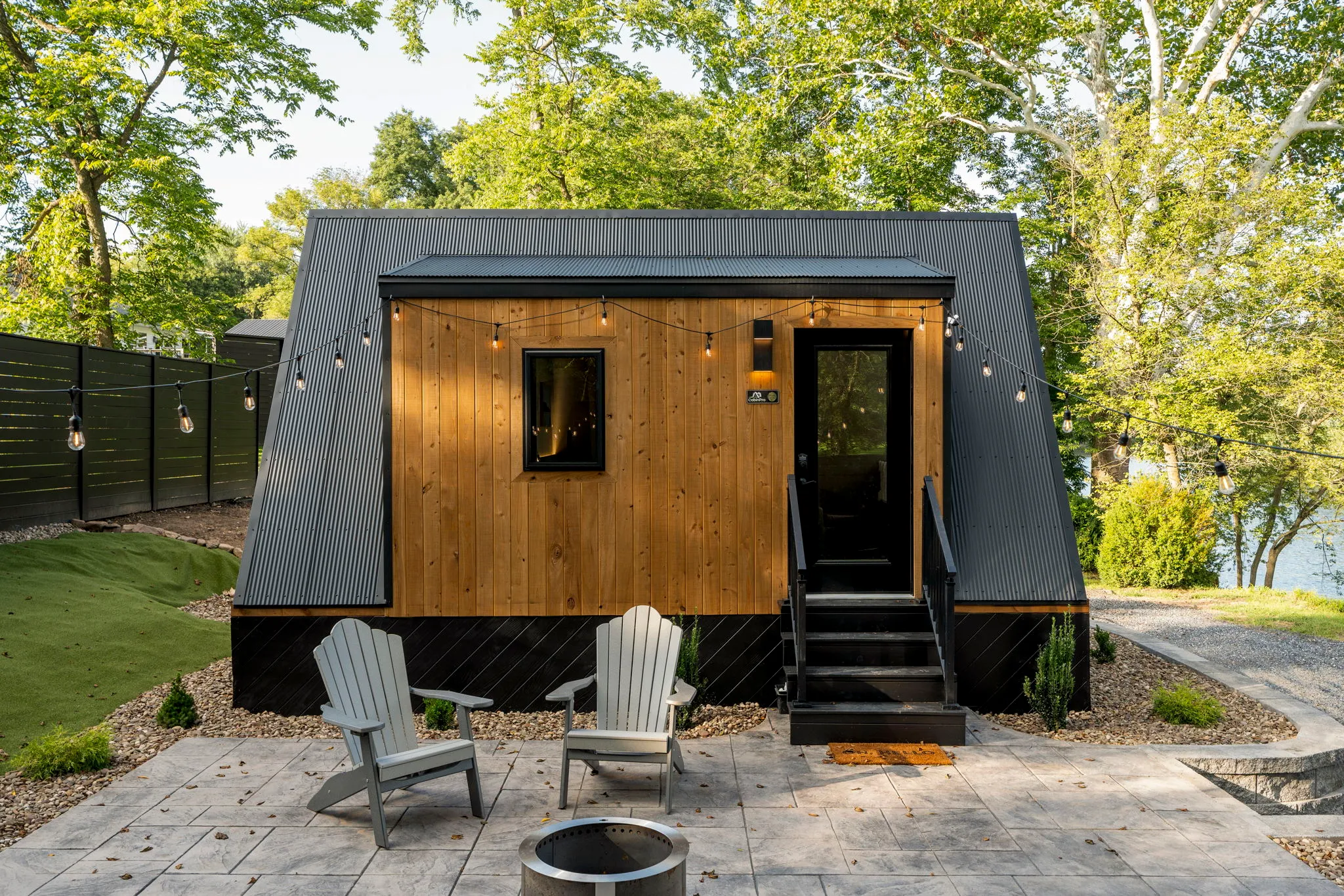 Modern tiny cabin with warm wood siding, black trim, two Adirondack chairs and a fire pit on a patio, surrounded by trees.