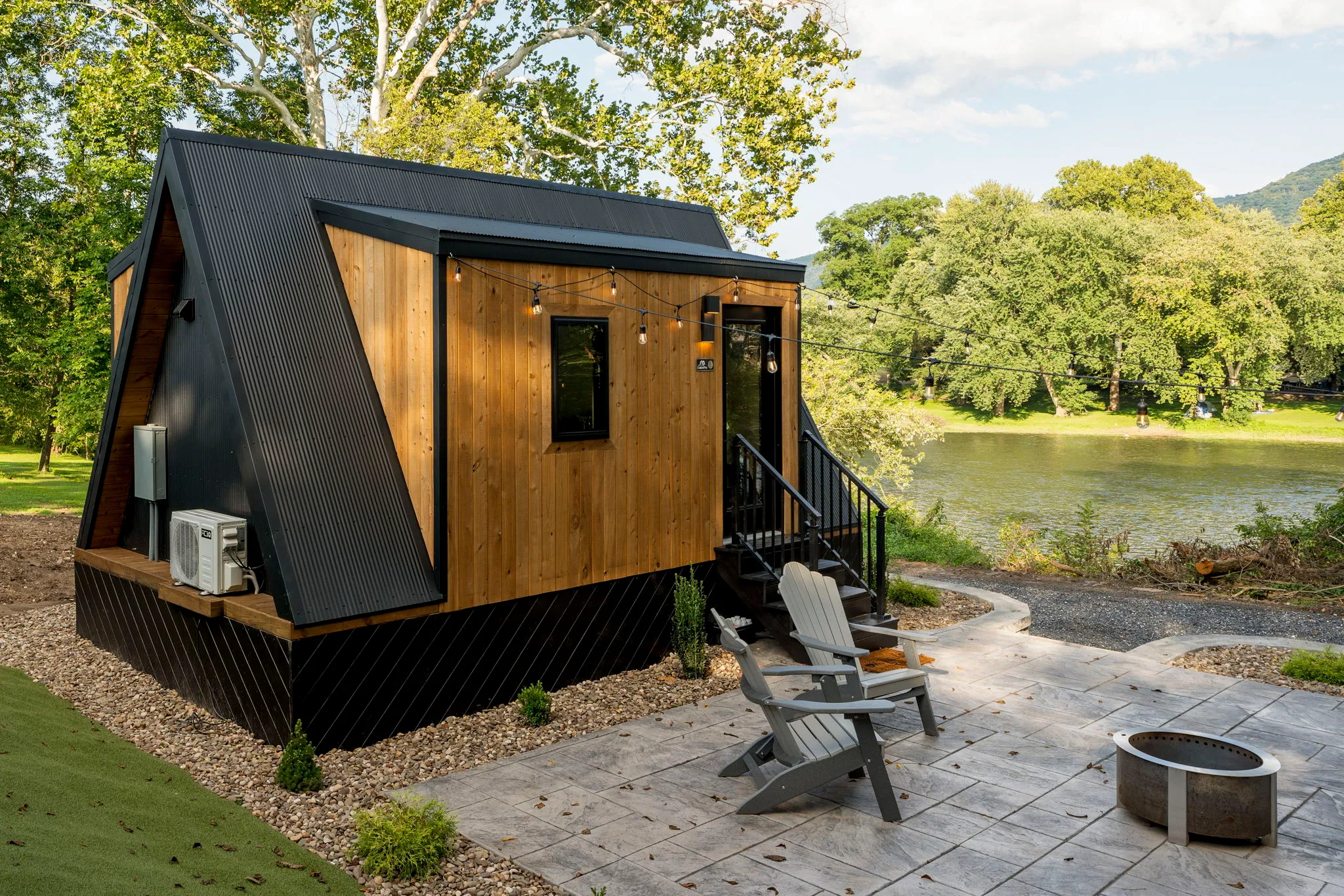 Modern black-and-wood tiny cabin with patio chairs and fire pit beside a peaceful lake, surrounded by trees and mountain views.