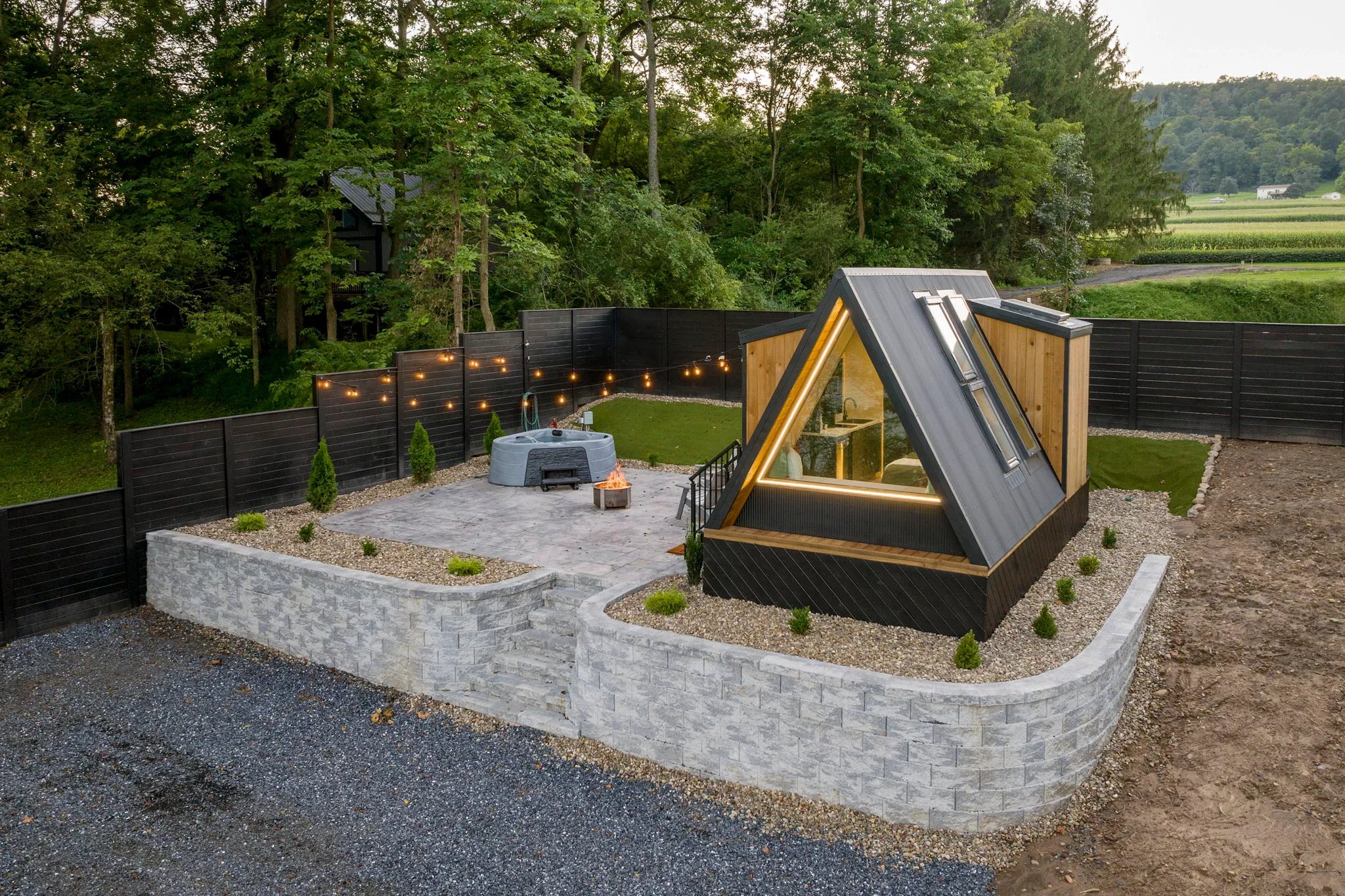 Modern A-frame garden cabin with glass front, landscaped stone terrace, gravel path, warm string lights, dark fence, and peaceful wooded farmland.