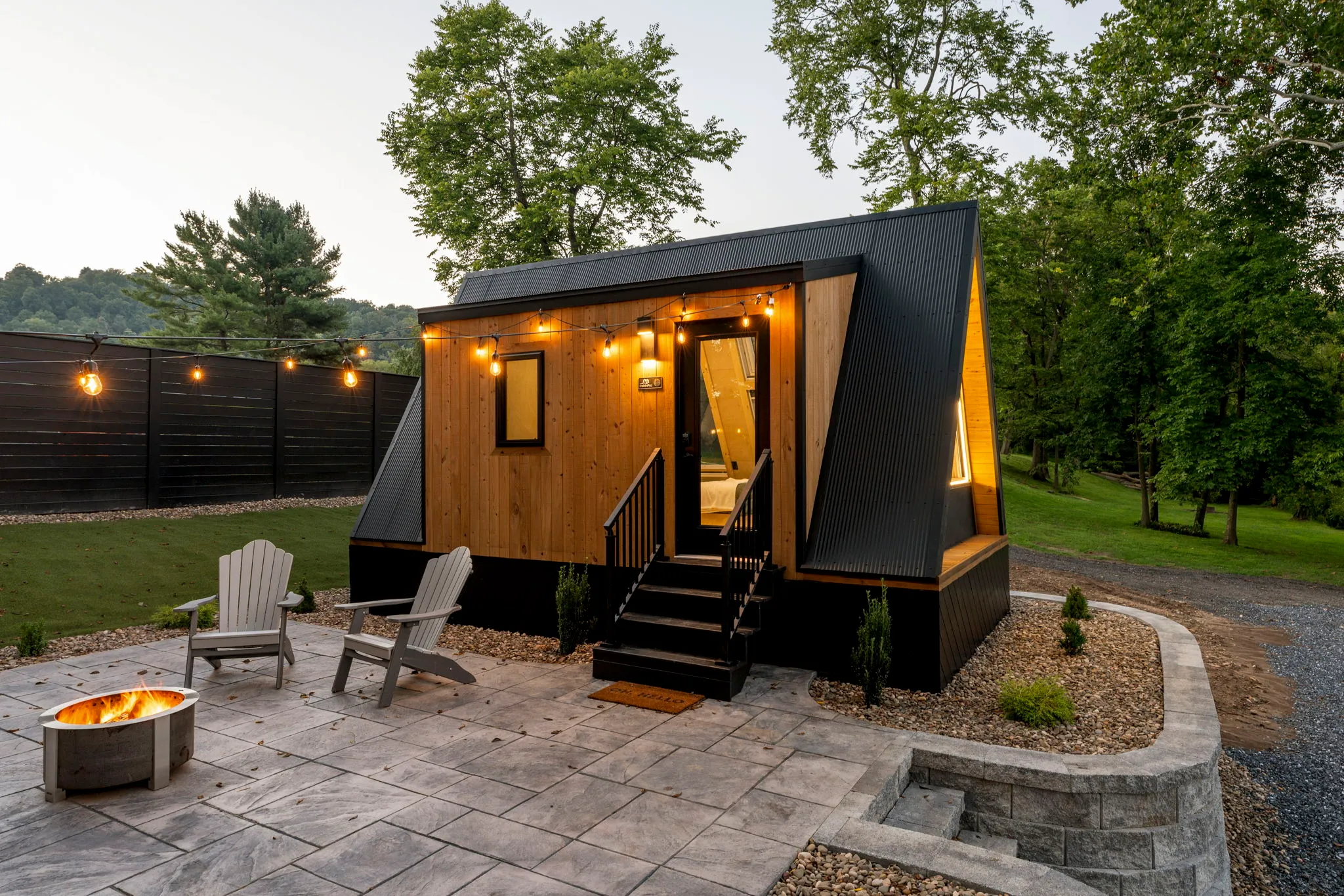 Modern tiny cabin with warm lights, patio, Adirondack chairs, and fire pit, surrounded by trees; cozy, inviting evening retreat.