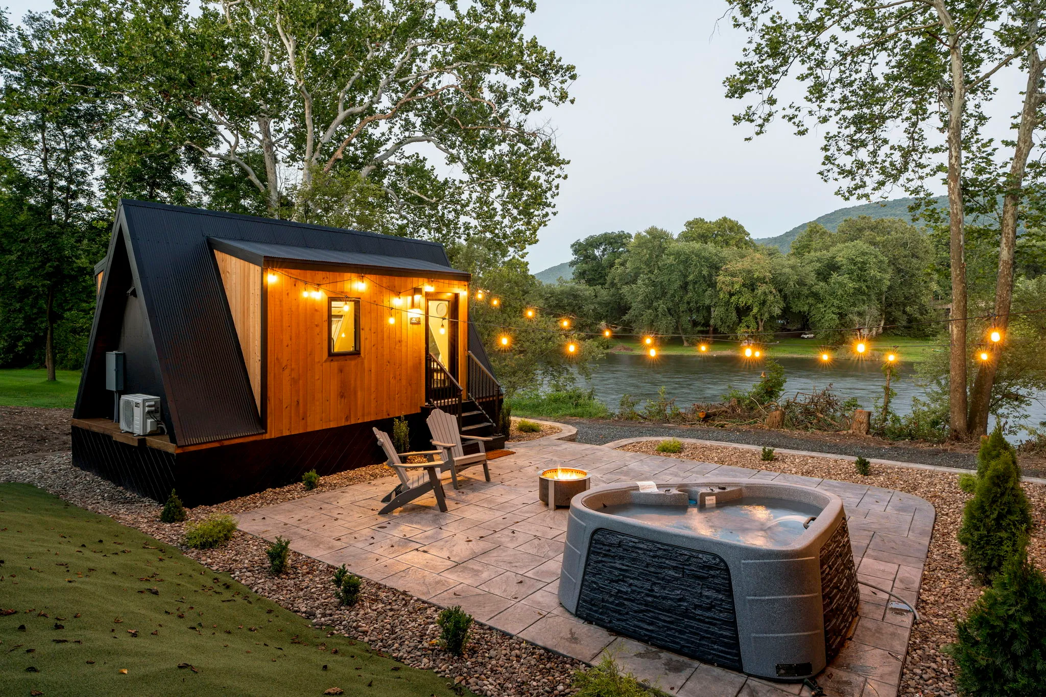 Cozy tiny cabin with warm lights, patio chairs, and bubbling hot tub beside a tranquil lake, surrounded by trees at dusk; peaceful retreat.