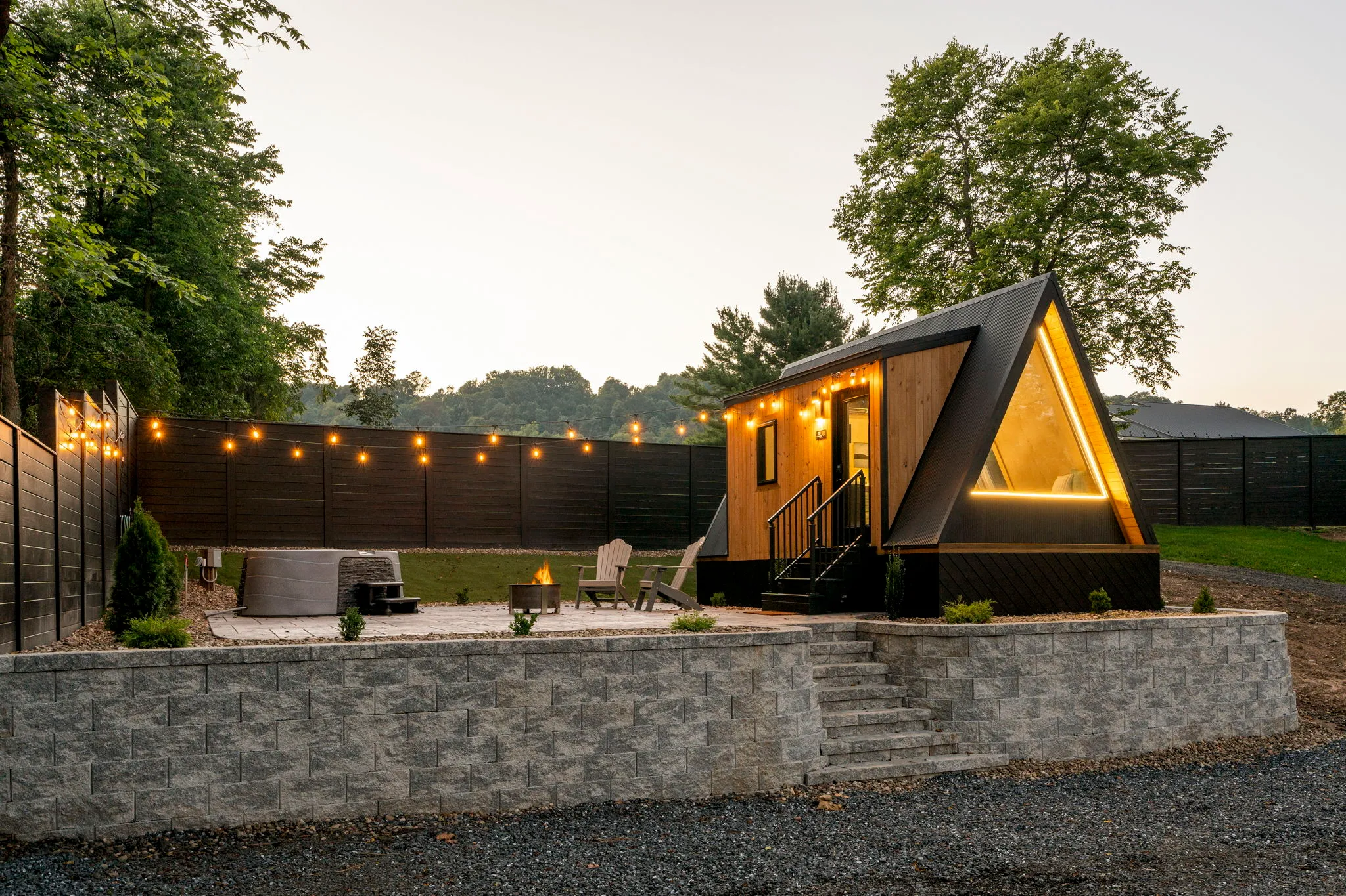 Modern A-frame cabin with glowing windows sits in a backyard patio, surrounded by string lights, trees, and a stone retaining wall.