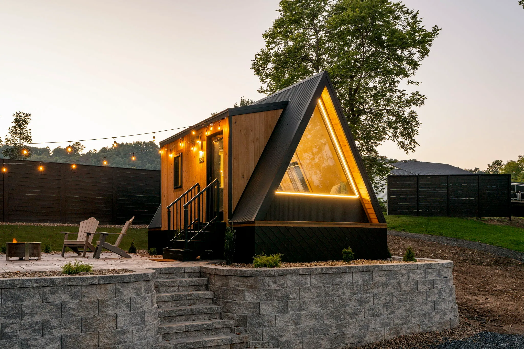A cozy modern A-frame tiny cabin glows warmly at dusk, with a triangular window, string lights, outdoor chairs, and a stone wall.