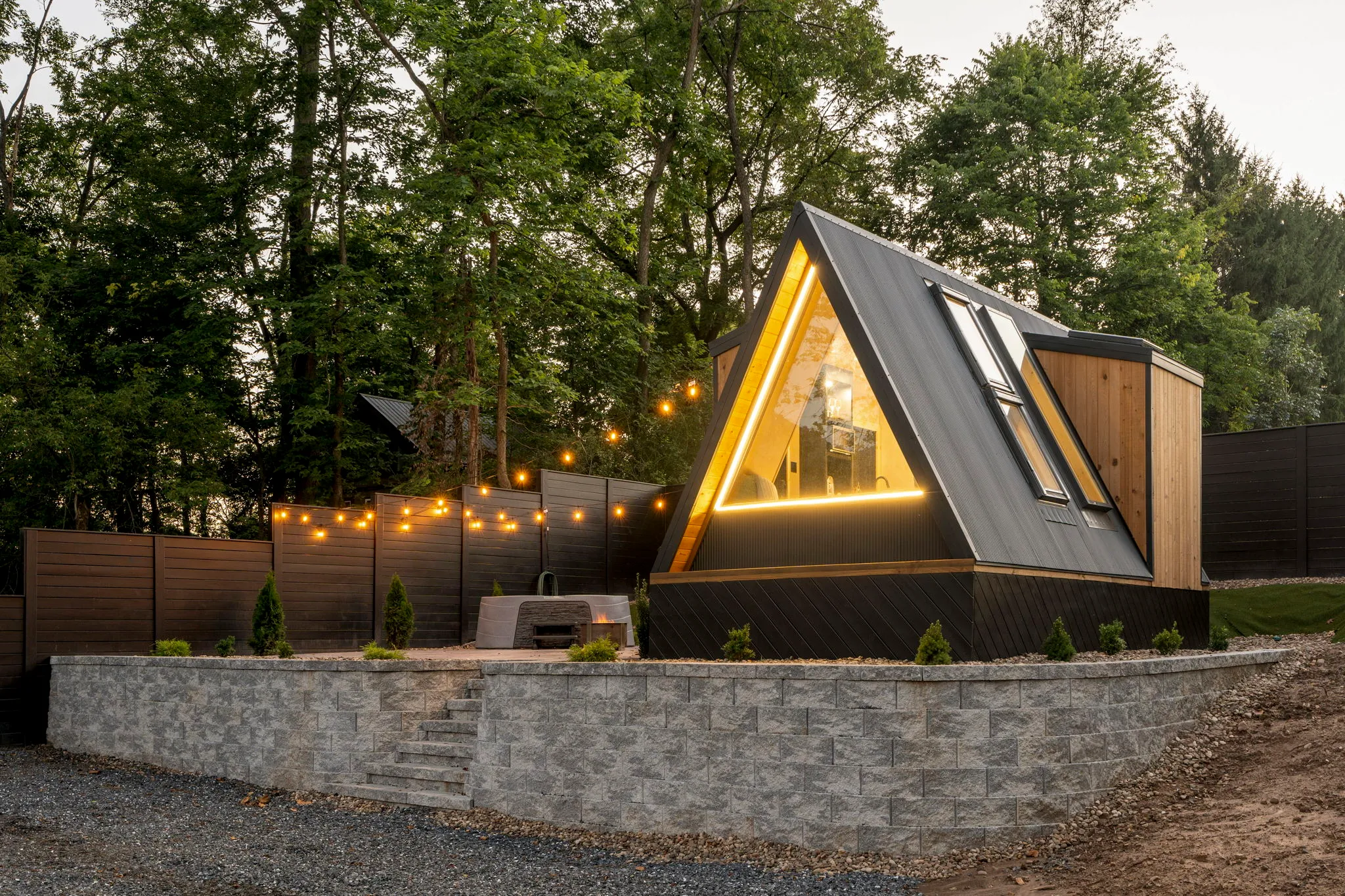 A cozy A-frame cabin glows warmly at dusk in a forest setting, with string lights, a timber fence, gravel path, and stone retaining wall.