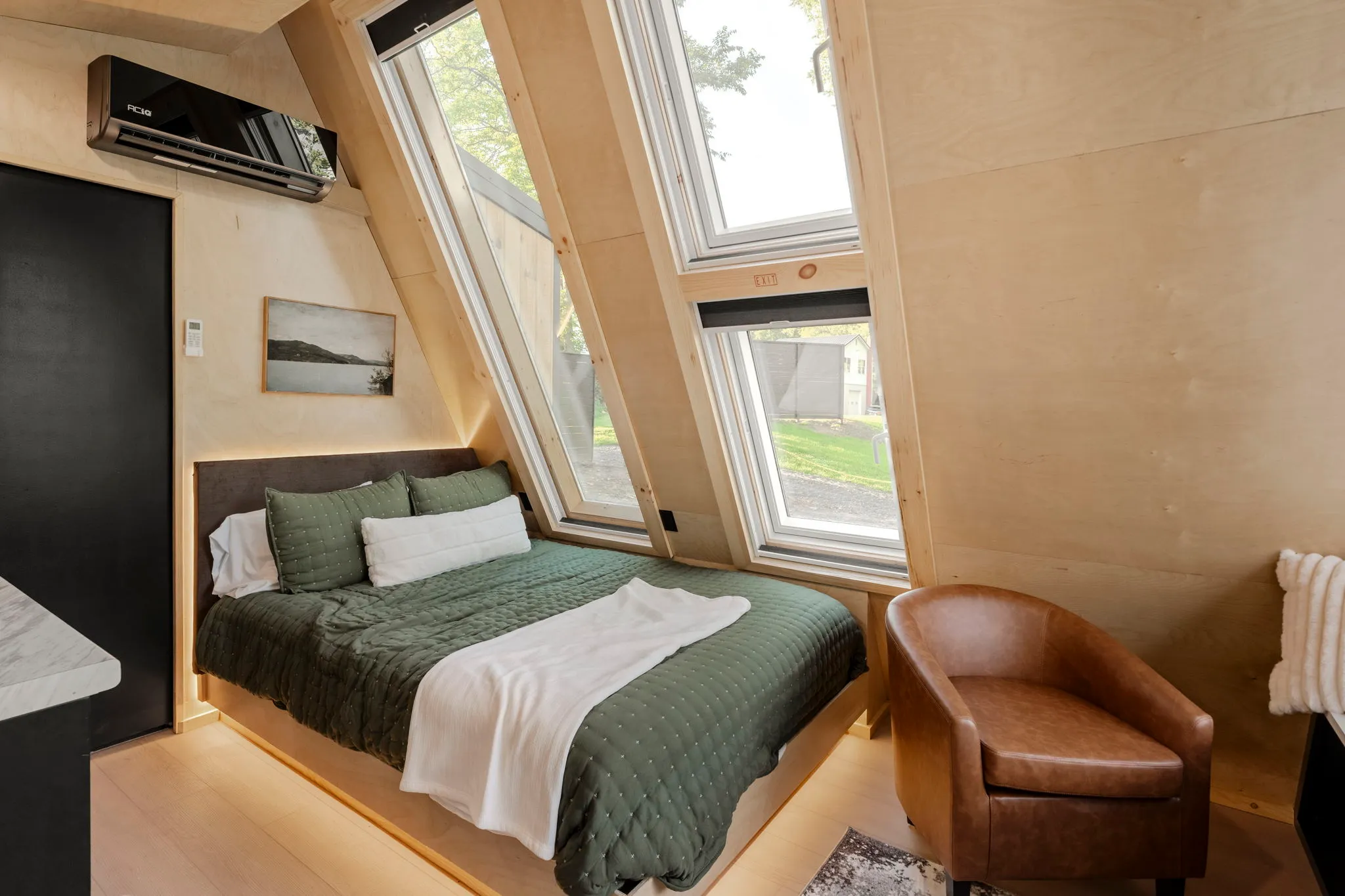 Cozy wood-paneled bedroom with a neatly made bed, skylight windows, green quilt, leather chair, and bright natural light.