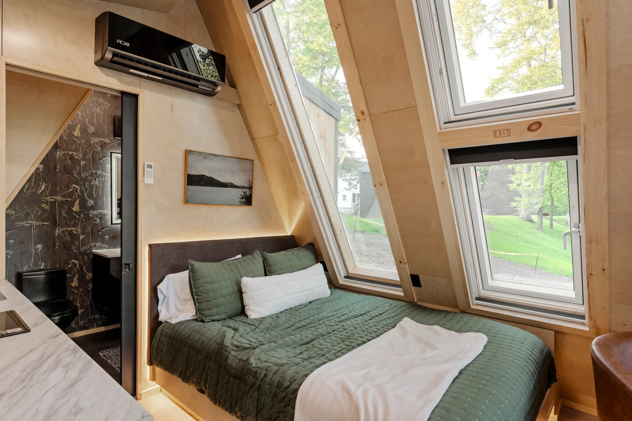 Cozy modern cabin bedroom with a green-covered bed, skylights, wood walls, and large windows overlooking a peaceful forested yard.