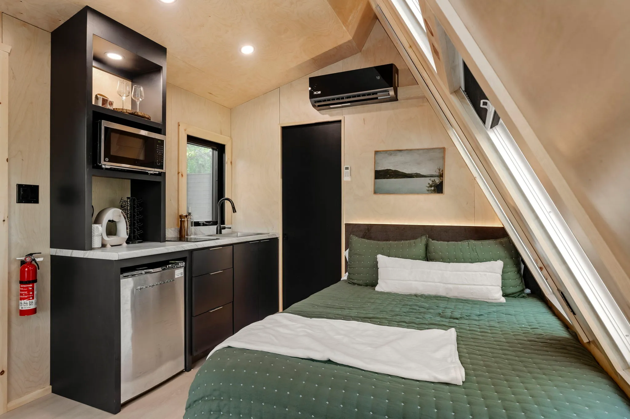 Cozy tiny cabin bedroom with a green-covered bed, compact kitchenette, black cabinets, skylights, and warm wood-paneled walls.