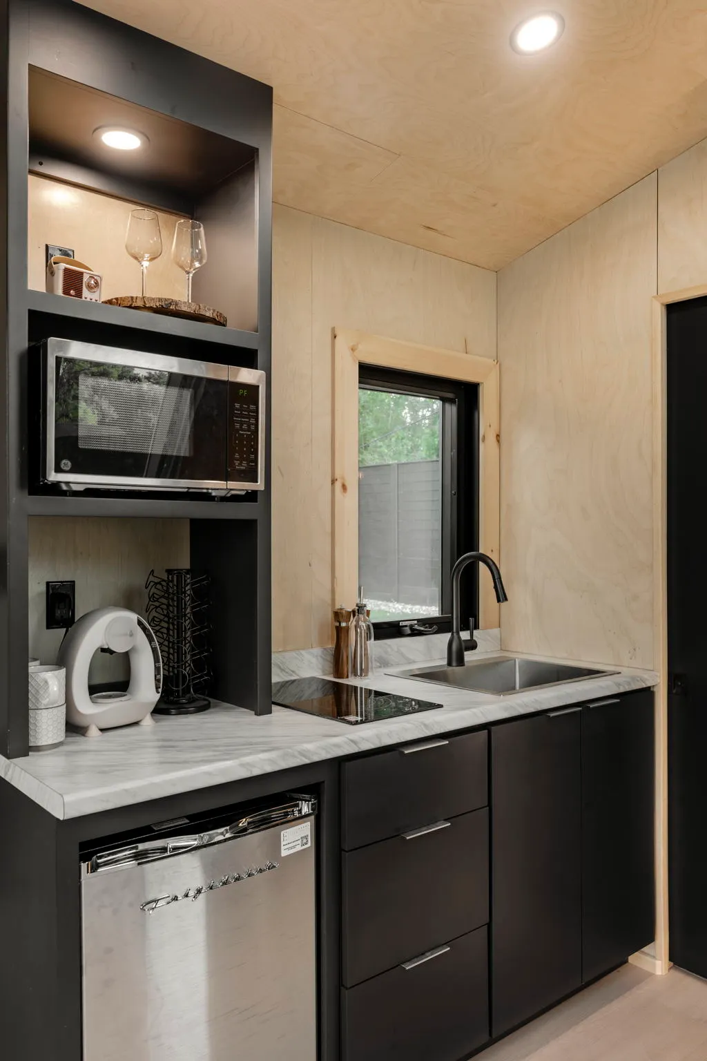 Compact modern kitchenette with black cabinets, stainless dishwasher, microwave, sink, open shelving, and warm wood walls in a cozy space.
