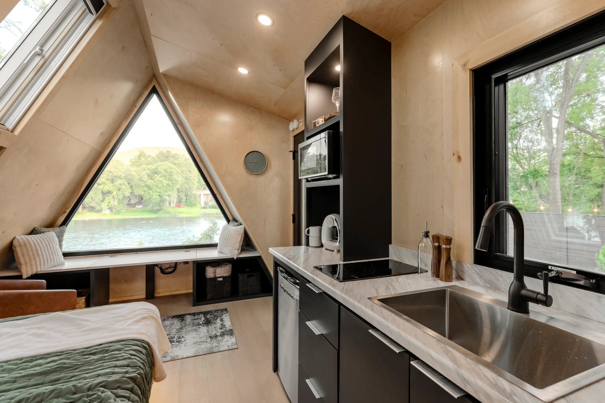 Cozy modern tiny home interior with kitchenette, triangular window overlooking a lake, sleeping nook, and warm wood finishes.