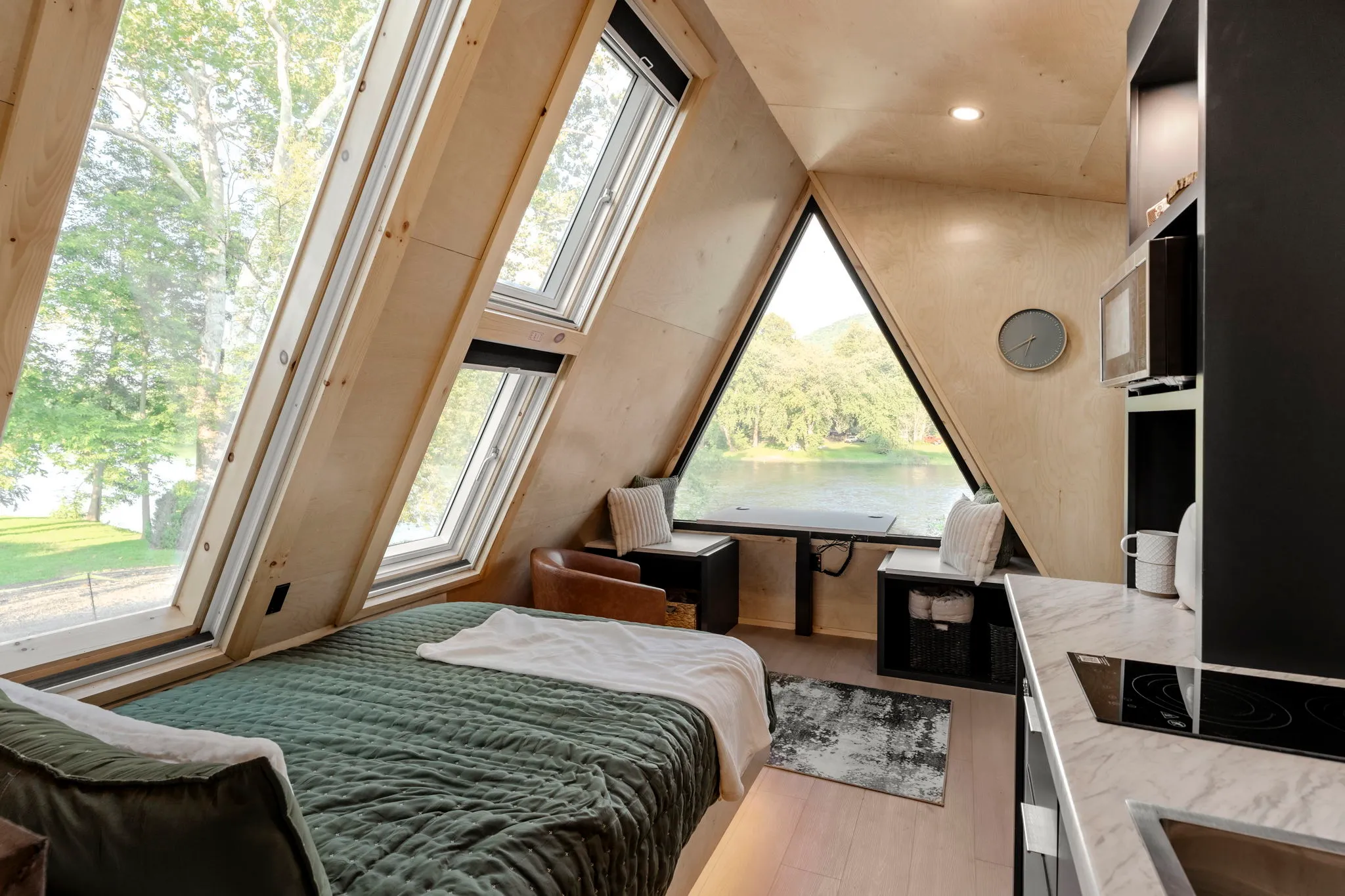 Cozy A-frame bedroom with a green-covered bed, skylights, kitchenette, and triangular window overlooking a peaceful lake and trees.