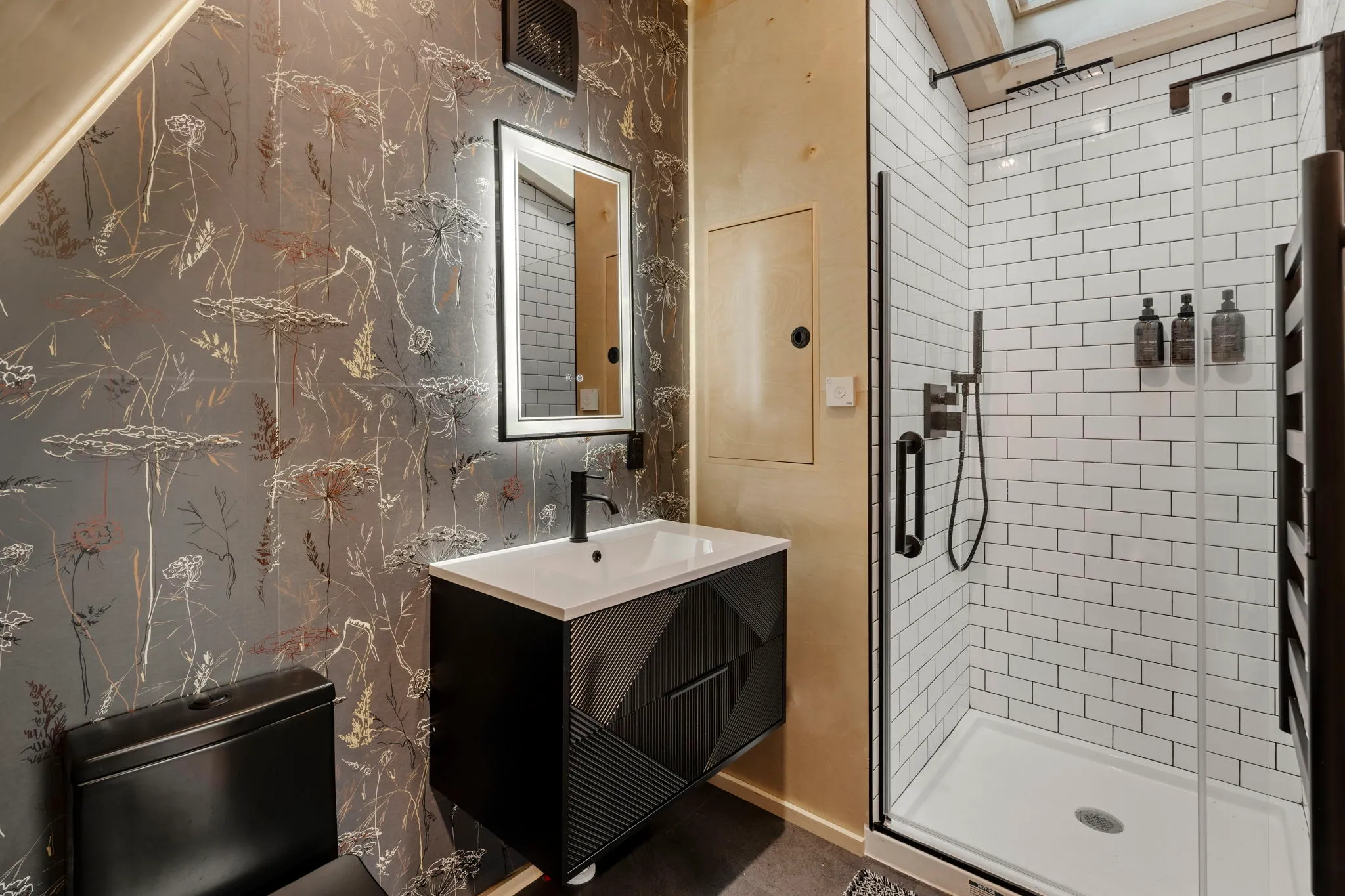 Modern, stylish bathroom with floral wallpaper, black vanity and toilet, mirrored cabinet, and glass-enclosed white subway-tile shower.