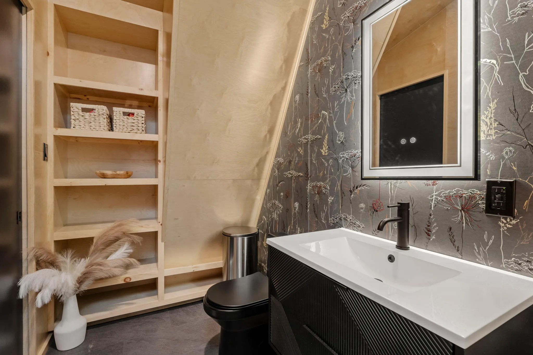 Stylish bathroom with black vanity, white sink, floral wallpaper, framed mirror, toilet, and built-in shelves with baskets and decor.
