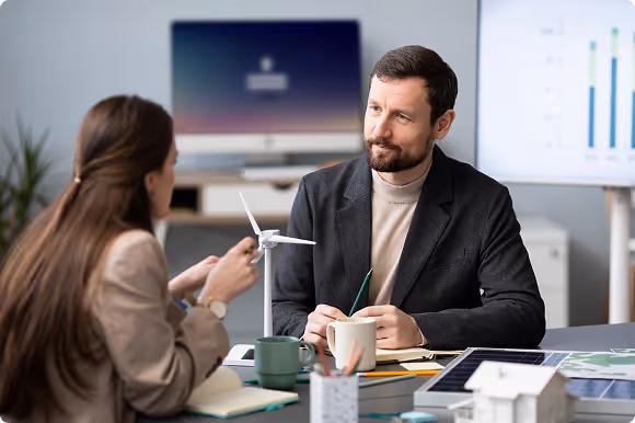 Two colleagues discussing a small wind turbine model at a desk with coffee cups, notebooks, and a tablet, with charts displayed on screens in the background.