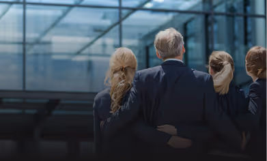 Four businesspeople with backs facing the camera, two men and two women, standing closely together in front of a modern glass building.