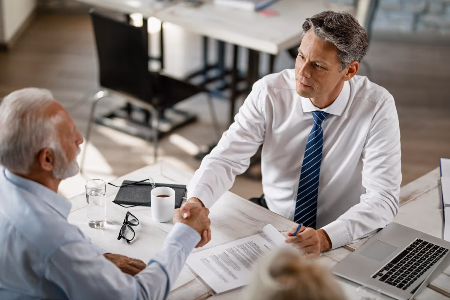 Two men in a business setting shaking hands across a table with documents and a laptop.