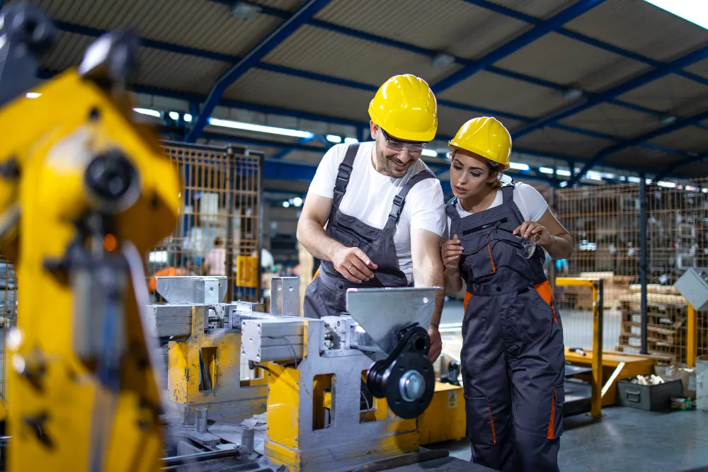 Two factory workers wearing yellow helmets and gray overalls inspecting machinery in an industrial workshop.