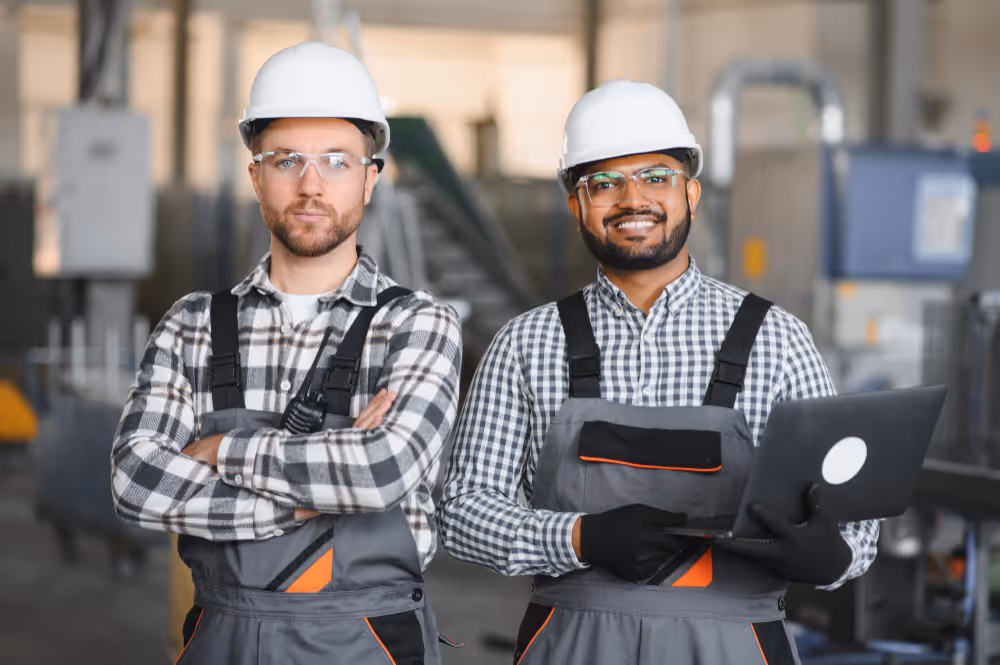 Two male factory workers wearing safety helmets and overalls, one with folded arms and the other holding a laptop, standing in an industrial setting.