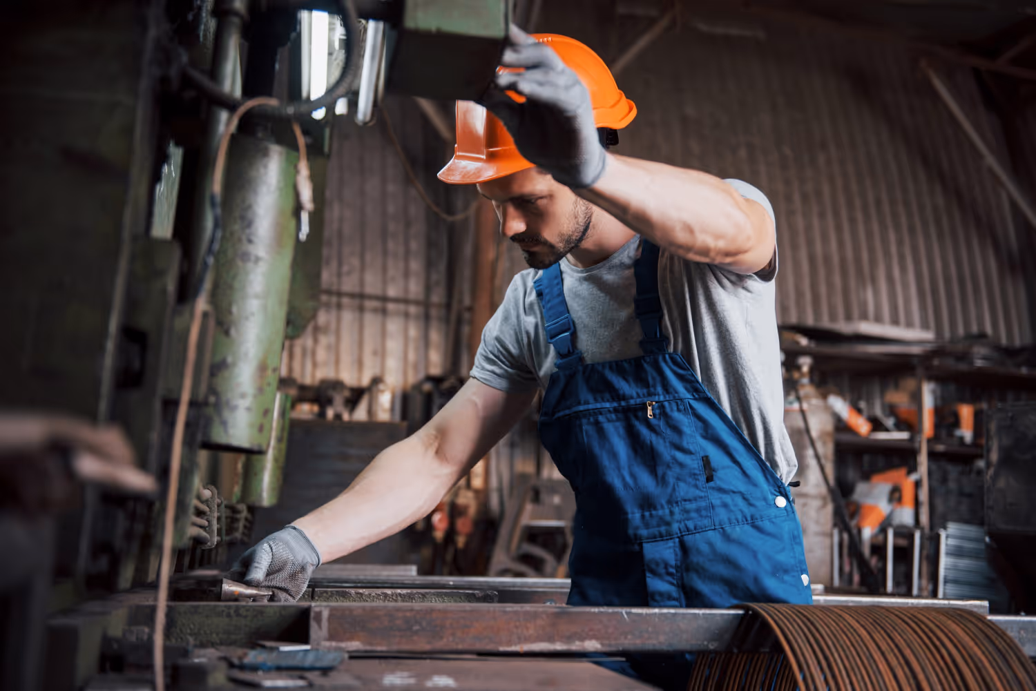 Worker wearing an orange hard hat and blue overalls operating industrial metalworking machinery in a workshop.