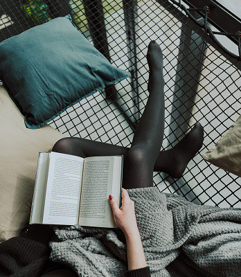 Osoba w czarnych rajstopach czytająca książkę na huśtawce z poduszkami i kocem.