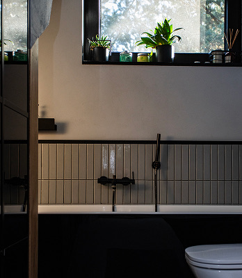 Nowoczesna łazienka z wanną, czarną armaturą i roślinami na parapecie podświetlonym naturalnym światłem.