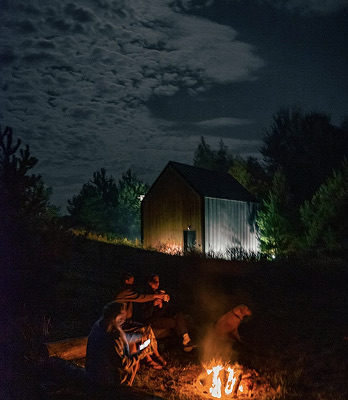 Trzech mężczyzn siedzi przy ognisku na zewnątrz nocą, w tle drewniany domek otoczony ciemnymi drzewami i pochmurne niebo.