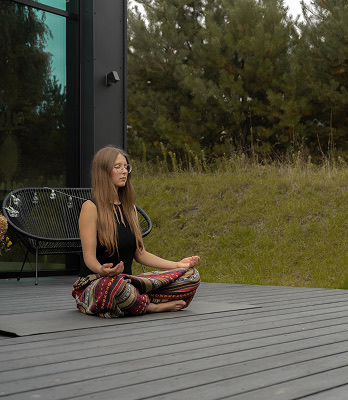 Kobieta w kolorowych spodniach siedzi ze skrzyżowanymi nogami na tarasie na zewnątrz, medytując z zamkniętymi oczami.