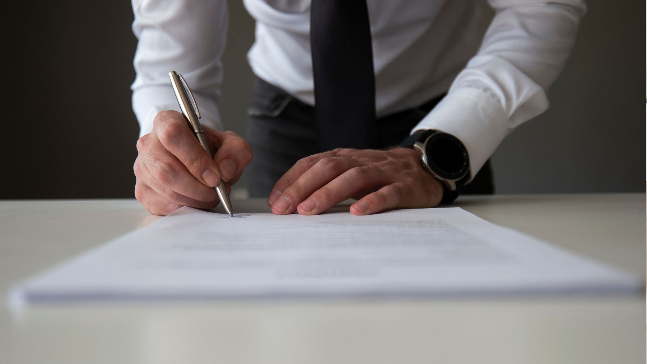 Osoba v bílé košili a černé kravatě podepisuje dokument stříbrným perem na stole.