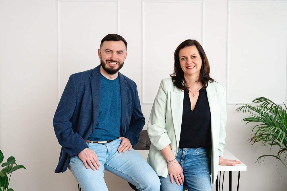 Dva usměvaví dospělí, muž a žena, sedící a opírající se o stůl v moderní červeno-bílé místnosti s rostlinami.