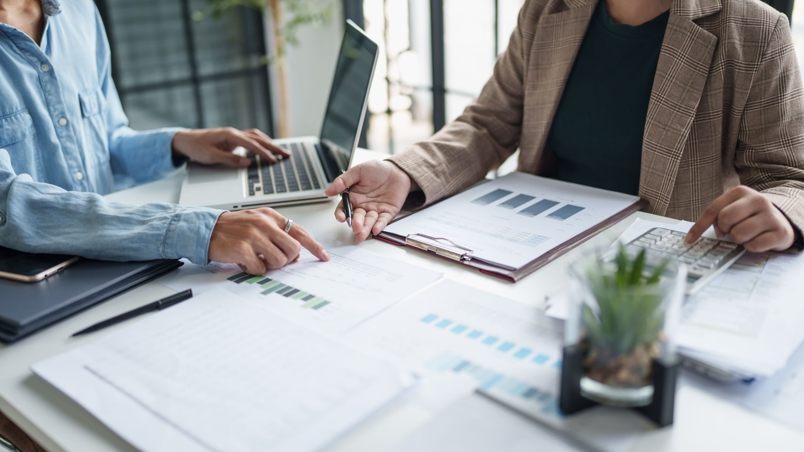 Dva lidé spolupracují u stolu s dokumenty, grafy, notebookem a kalkulačkou.