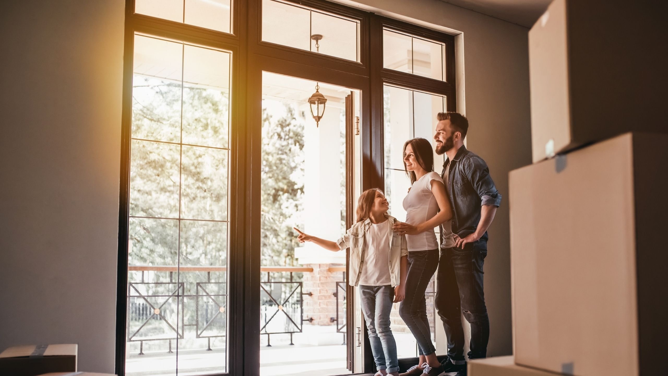 Rodina s malou dcerou stojí u otevřených dveří na terasu v novém domě s krabicemi po přestěhování.