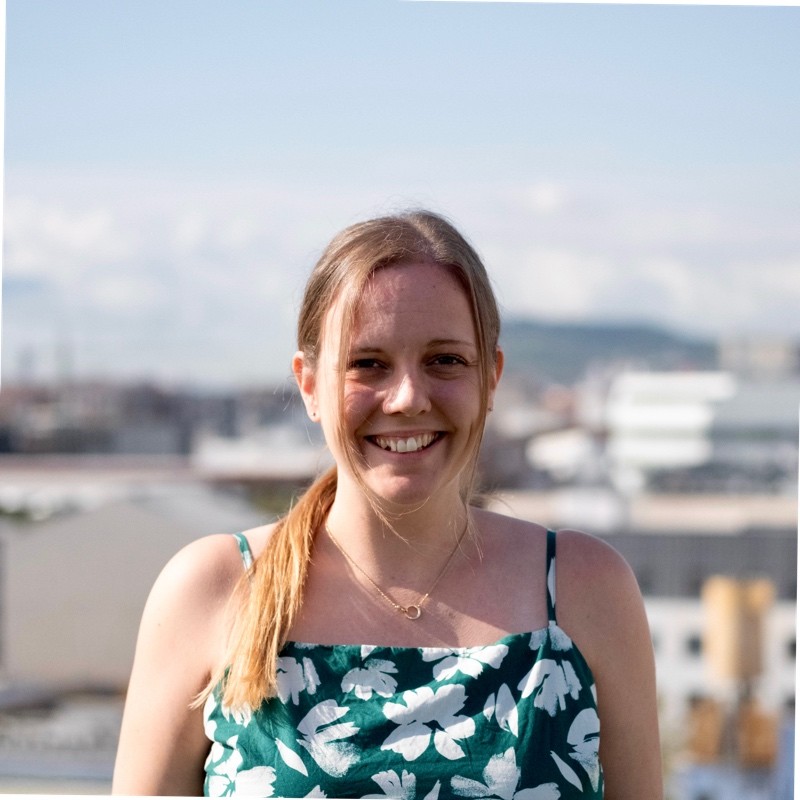 Usměvavá žena s dlouhými světlými vlasy v culíku, oblečená v zelených květovaných šatech, stojící venku s rozostřeným městským pozadím.