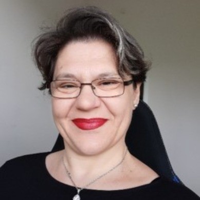 Usmívající se žena se světlými prameny ve vlasech, brýlemi a červenou rtěnkou, na šedém pozadí.