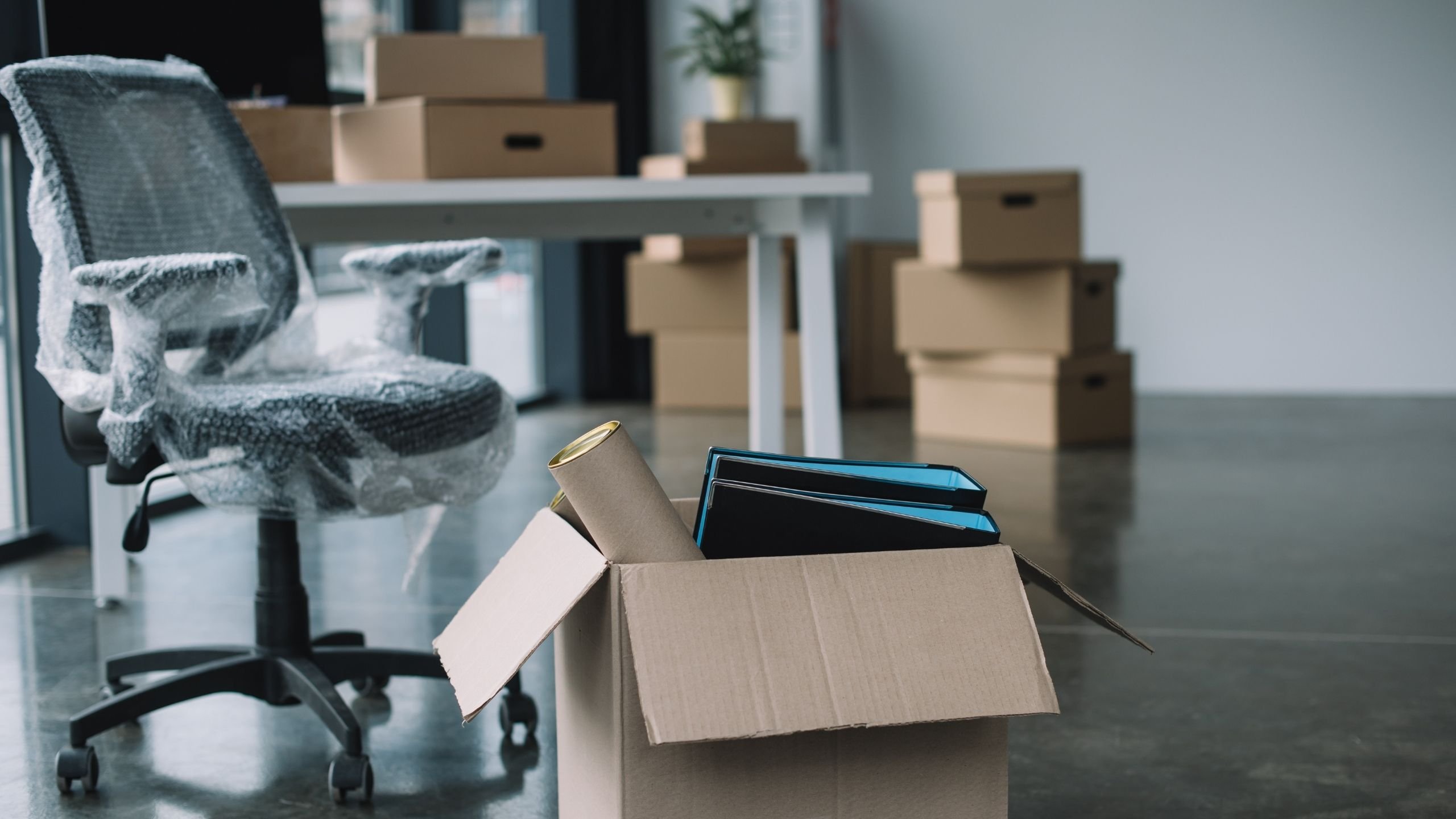 Kancelářská židle zabalená v bublinkové fólii vedle rozbalené lepenkové krabice s pořadači a tubusem v prázdné kanceláři s hromadami krabic v pozadí.
