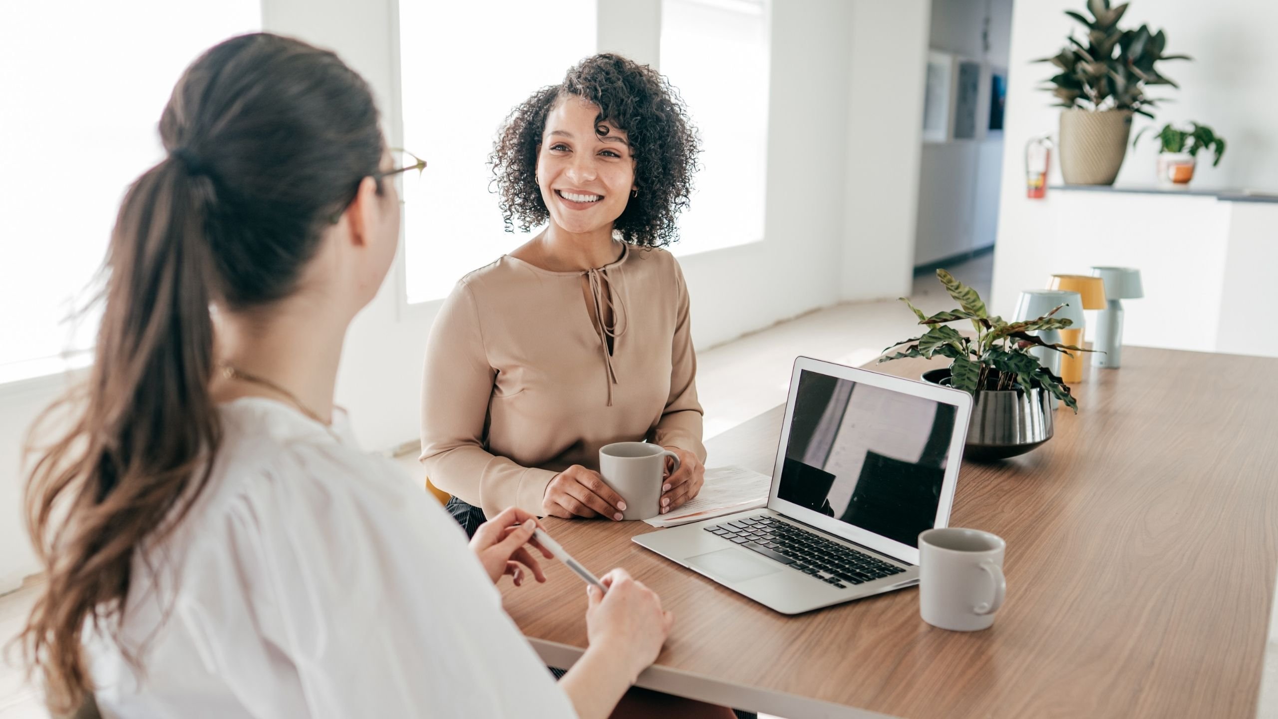 Two women are sitting at a table, one of them is holding a mug and smiling during the conversation, there is an open laptop on the table and another a mug.