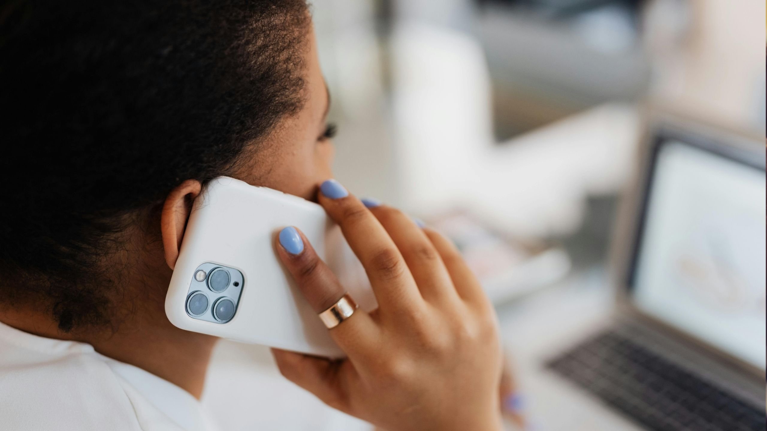 Osoba držící bílý smartphone u ucha při telefonování, na pozadí rozostřený notebook.