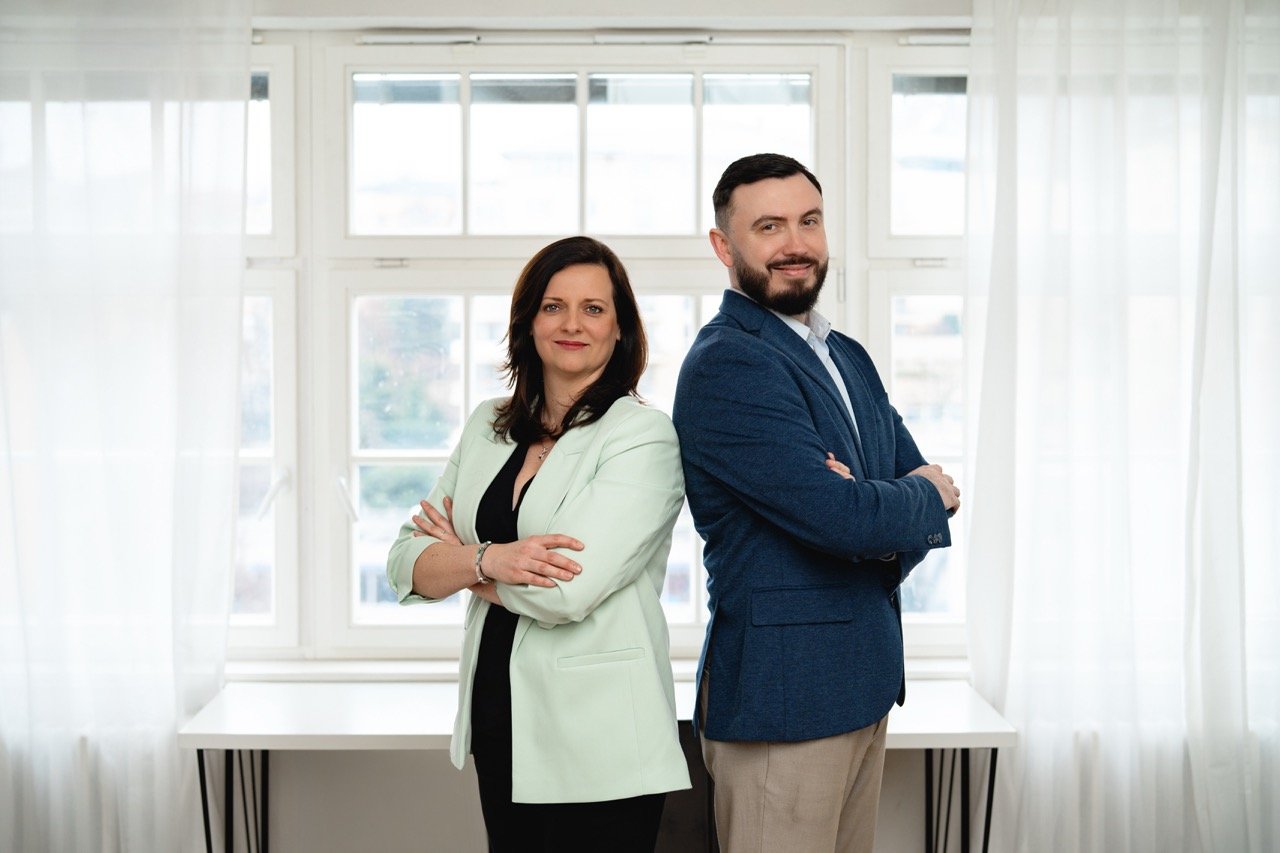 Muž a žena stojící zády k sobě s překříženýma rukama a usmívající se v jasné místnosti s oknem a bílými závěsy.