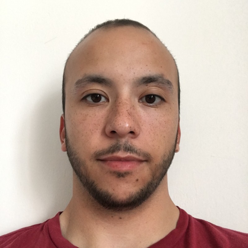 Muž s krátkými vlasy, jemným vousem a pihami na tváři, oblečený v červeném tričku, pózuje před bílou stěnou.