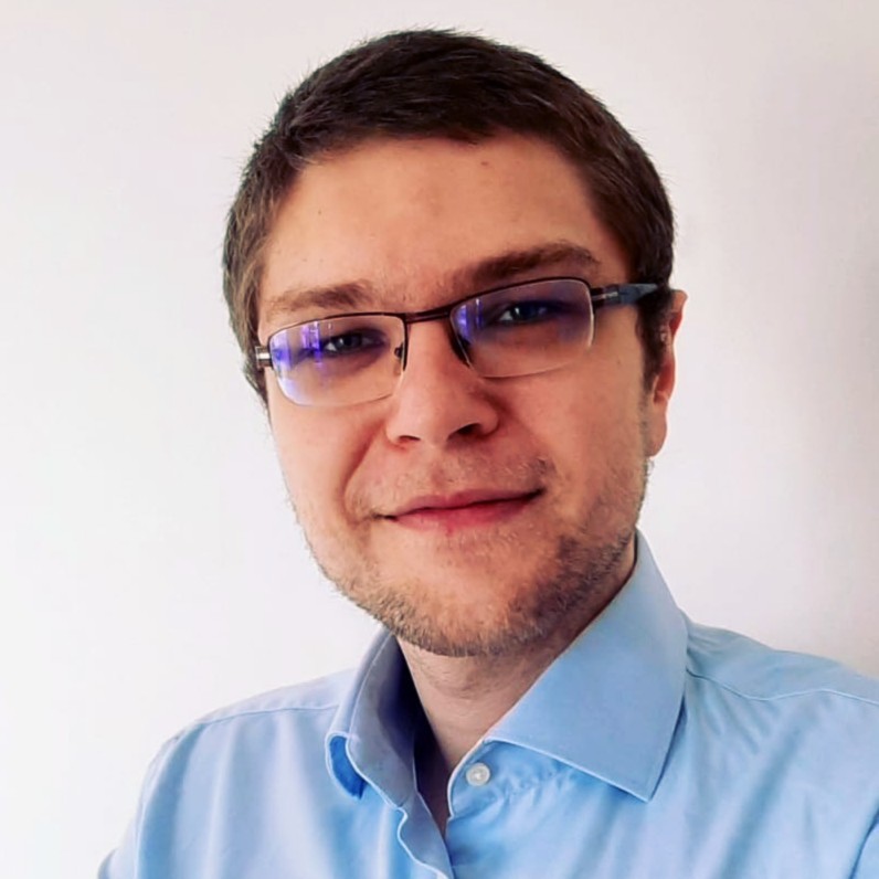 Muž s krátkými hnědými vlasy, brýlemi a modrou košilí se usmívá do kamery na světlém pozadí.