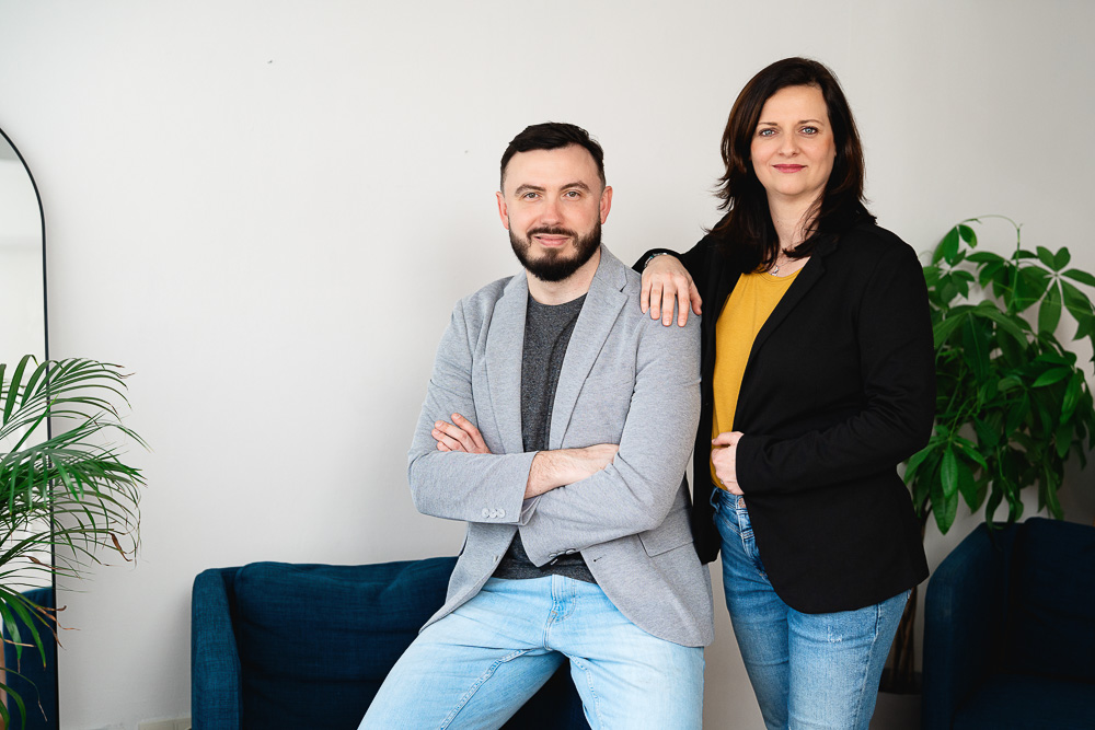 Muž v šedém saku s překříženýma rukama sedí na modré pohovce, zatímco žena v černém saku a žlutém tričku stojí vedle něj s rukou na jeho rameni, oba se usmívají v interiéru s bílou stěnou a zelenými rostlinami.