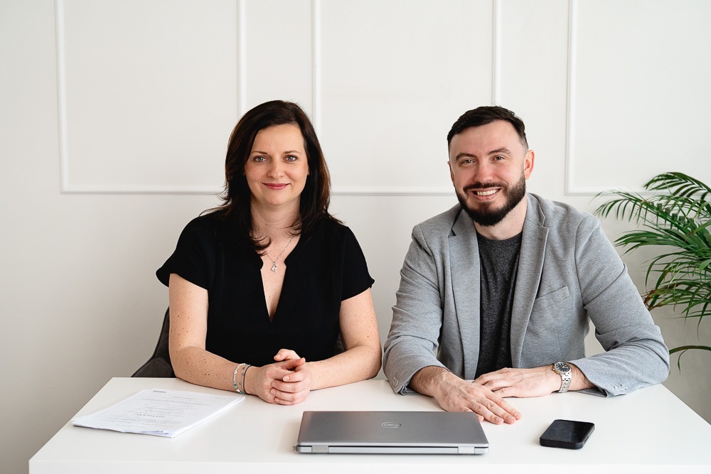 Muž a žena sedí za bílým stolem s notebookem, telefonem a dokumenty, oba se usmívají.